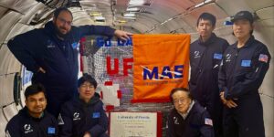 A group of six individuals in matching uniforms poses inside an aircraft, with banners from the University of Florida and MAE pinned on equipment.