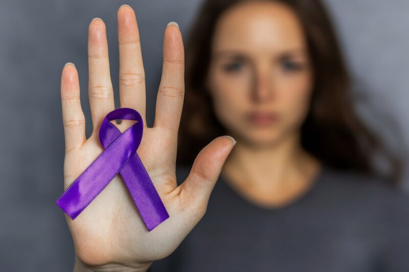 A person holds their open palm forward, displaying a purple awareness ribbon against a blurred background, symbolizing support for a specific cause.