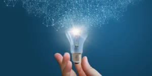 A hand holds a glowing light bulb against a blue background, with a network of glowing connections above, symbolizing innovation and ideas.