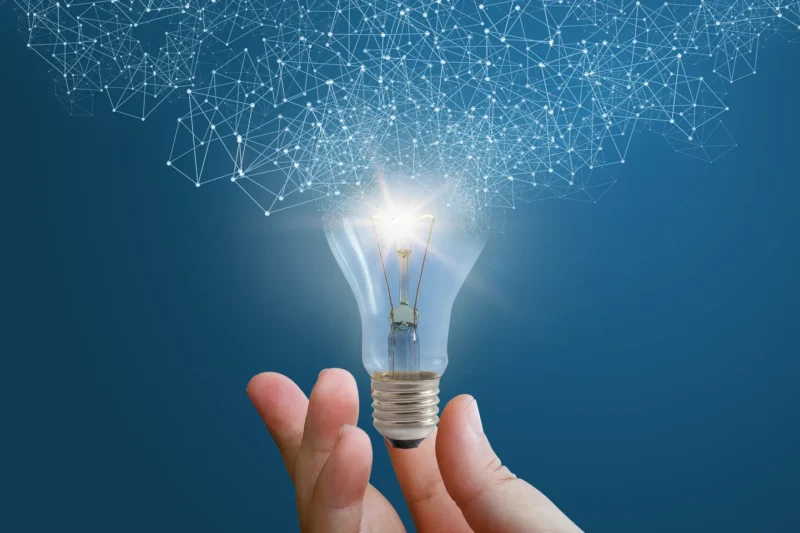 A hand holds a glowing light bulb against a blue background, with a network of glowing connections above, symbolizing innovation and ideas.