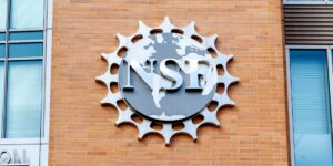 A metal National Science Foundation (NSF) logo is mounted on a brick wall, featuring a world map and gear-like border design.