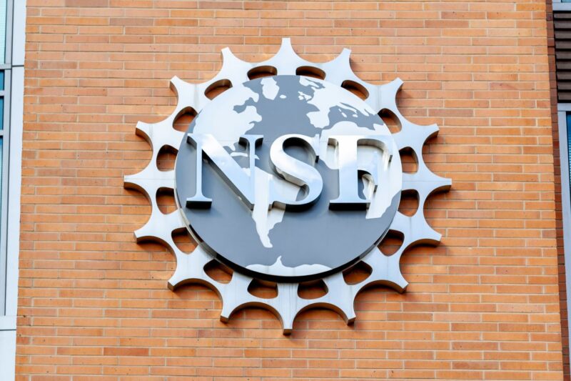 A metal National Science Foundation (NSF) logo is mounted on a brick wall, featuring a world map and gear-like border design.