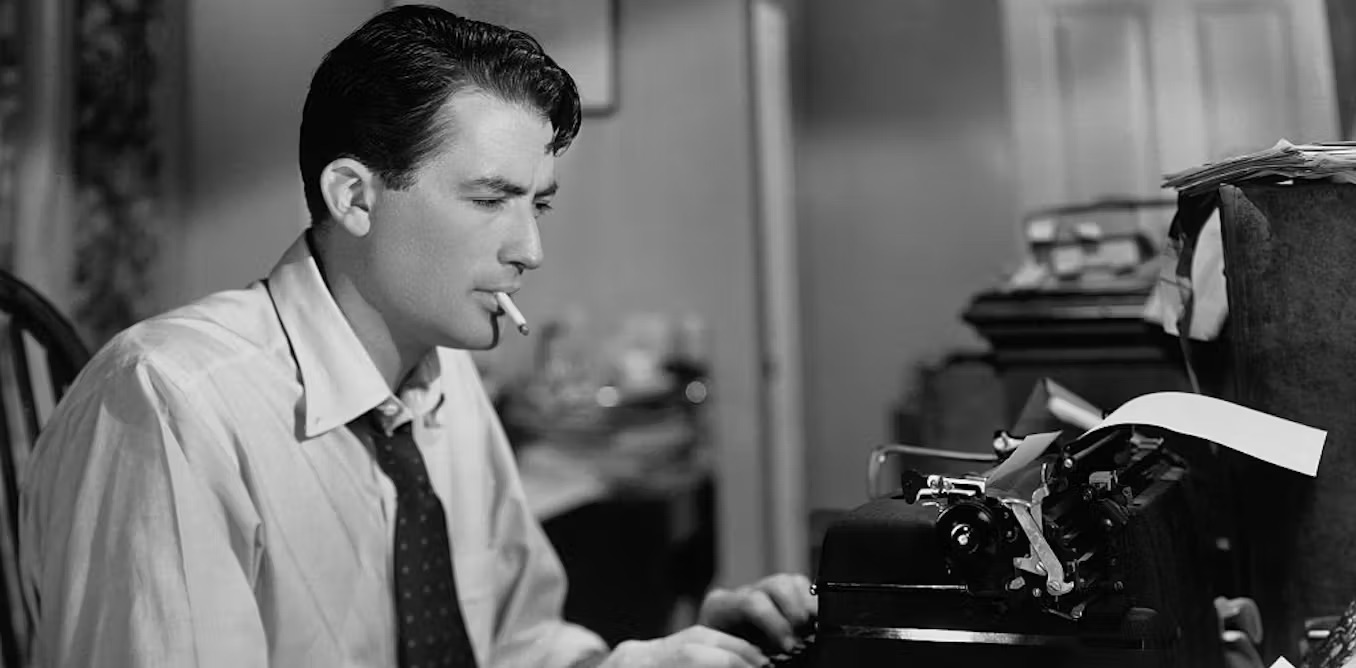 A black and white photo of a white male sitting at a typewriter with a cigarette in his mouth.