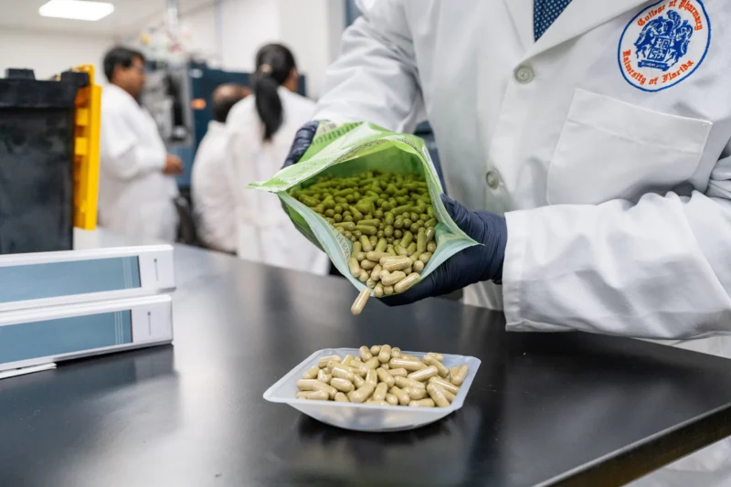 Lab technician in white coat and gloves pours green capsules into a tray in a pharmaceutical laboratory, with researchers blurred behind.
