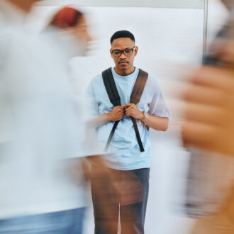 A young man with glasses and a backpack stands still in a hallway while blurred people walk past him, creating a sense of motion in the foreground.
