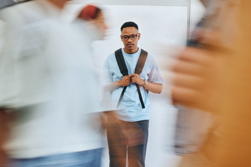 A young man with glasses and a backpack stands still in a hallway while blurred people walk past him, creating a sense of motion in the foreground.