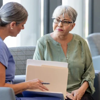 A healthcare worker in blue scrubs sits with an older woman in a green top, reviewing documents together in a bright, modern medical waiting area.