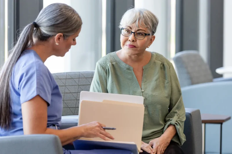 A healthcare worker in blue scrubs sits with an older woman in a green top, reviewing documents together in a bright, modern medical waiting area.
