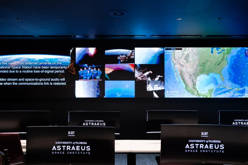 A dark mission control room with a large wall display showing space imagery and a map, with multiple computer monitors labeled Astraeus Space Institute.