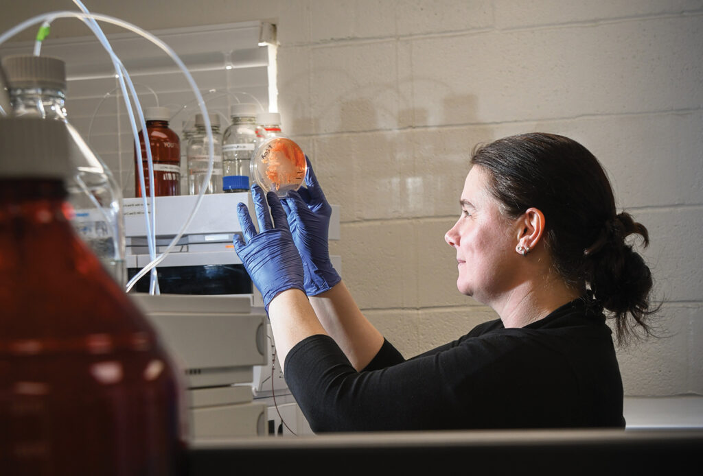 A photo of a woman wearing blue gloves and a black shirt in a chemistry lab holding a sample of a chemical.