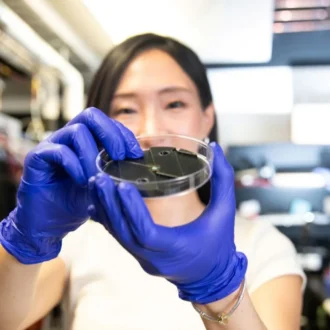 A scientist wearing blue nitrile gloves holds a clear petri dish containing a small, black, circular electronic component in a laboratory setting.