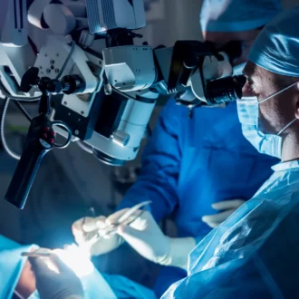 Two surgeons in blue scrubs, masks, and caps perform a delicate procedure using a specialized surgical microscope in a dimly lit operating room.