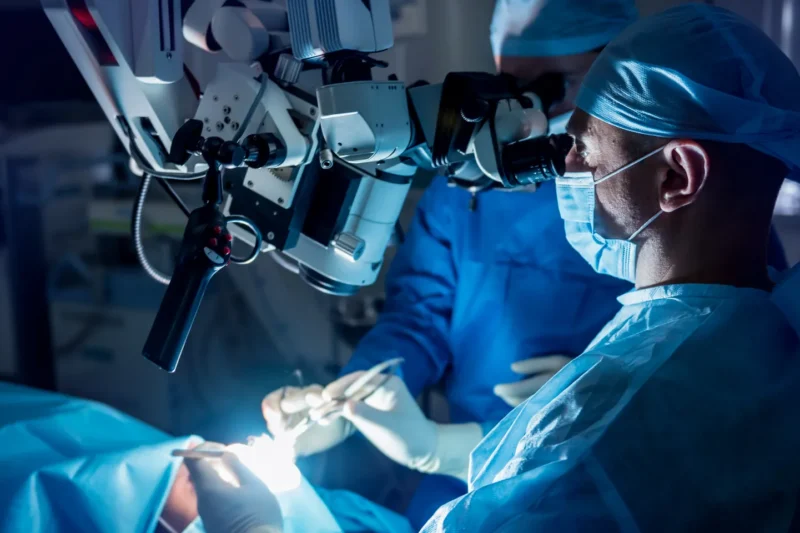 Two surgeons in blue scrubs, masks, and caps perform a delicate procedure using a specialized surgical microscope in a dimly lit operating room.